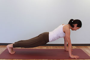 5.呼吸を止めて、もう一方の脚を後ろへ引き、両手と足の爪先で体重を支える。頭と胴を一直線に保ち（腕立て伏せの体勢）、両手の間の床一点を見る。